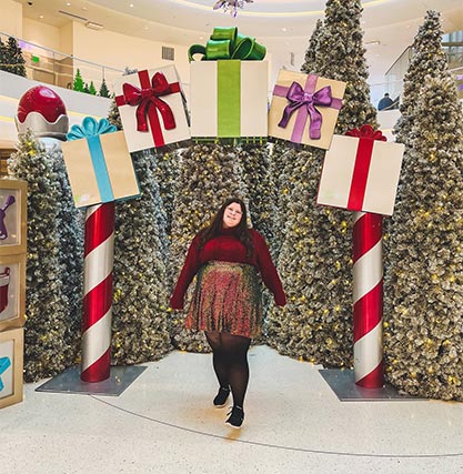 A photo of a woman posing for a picture in under an arch decorated with presents, in front of Christmas trees.