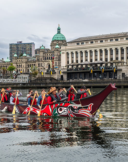 Songhees Nation indigenous peoples rowing a traditional boat on a river past a modern capitol building.